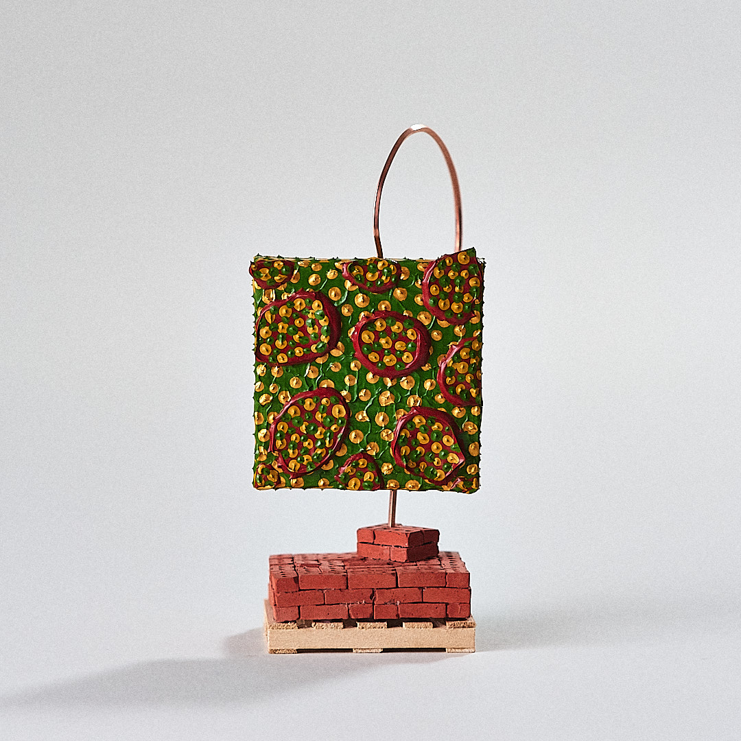 A green and gold painted form is held by curved copper wire over a miniature brick base.